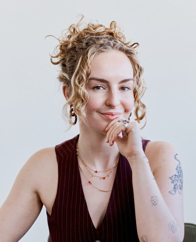 Rebecca, a blonde, white woman with blonde curly hair. She has tattoos and necklaces and is wearing a maroon waistcoat.