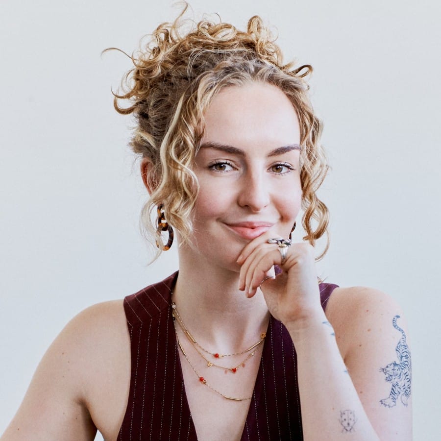 Rebecca, a blonde, white woman with blonde curly hair. She has tattoos and necklaces and is wearing a maroon waistcoat.