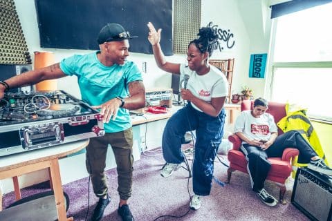 Brassic FM promotional image. One person on decks, one with a microphone dancing and one sat on an armchair scrolling on their phone