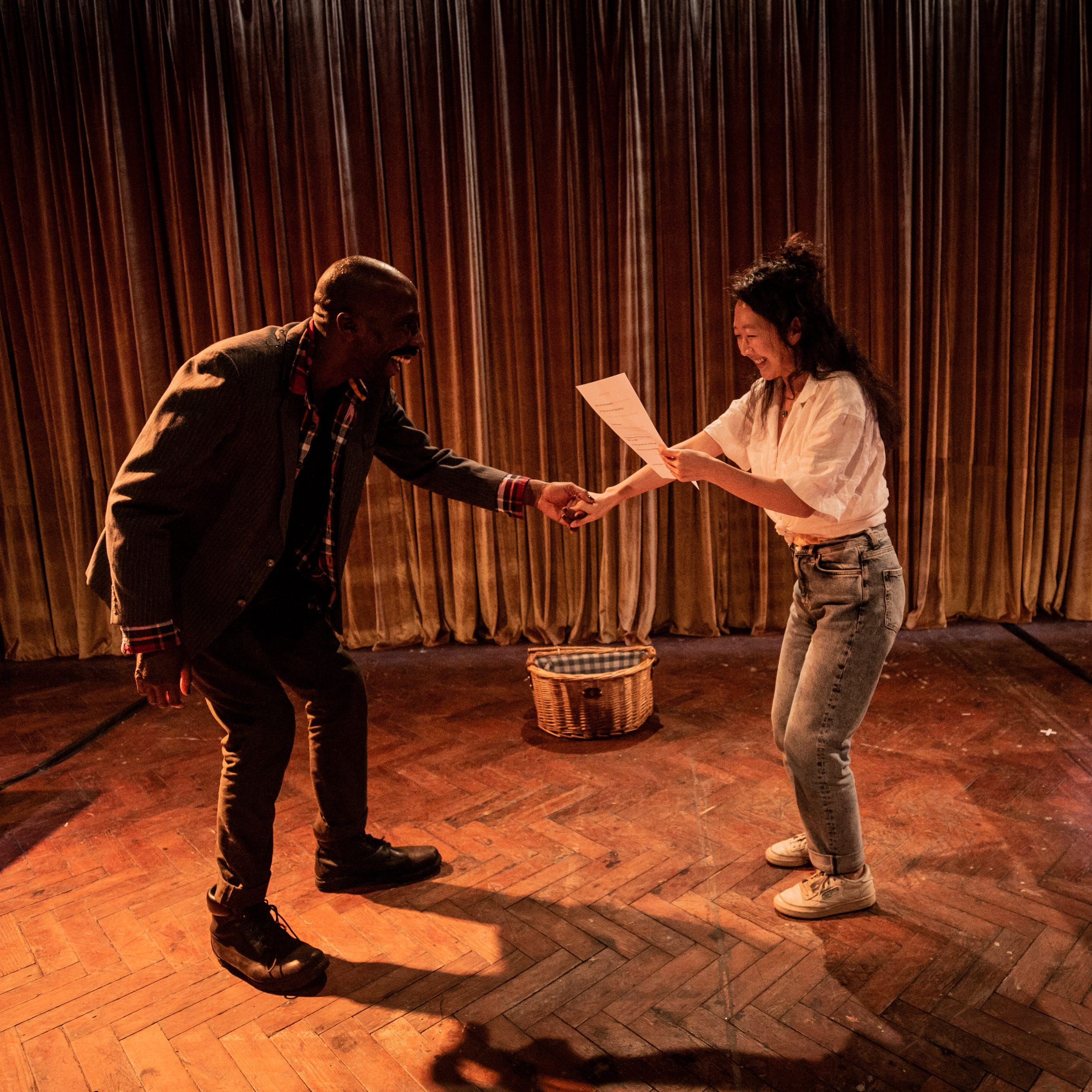 Two people, a man and a woman, share a lighthearted moment on a stage with wooden flooring. They stand near two white desks, lit by lamps. The man gestures with enthusiasm while the woman holds up a sheet of paper, smiling.