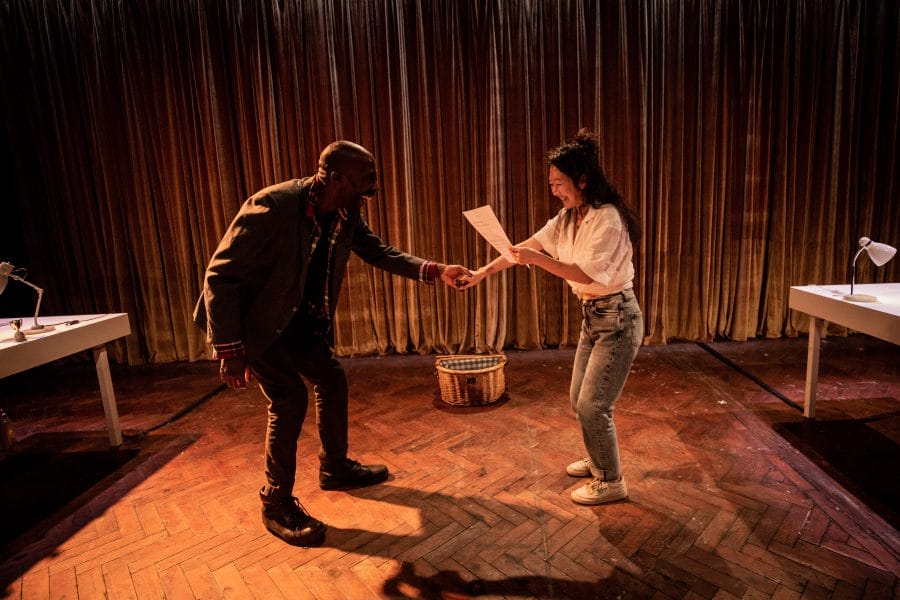 Two people, a man and a woman, share a lighthearted moment on a stage with wooden flooring. They stand near two white desks, lit by lamps. The man gestures with enthusiasm while the woman holds up a sheet of paper, smiling.
