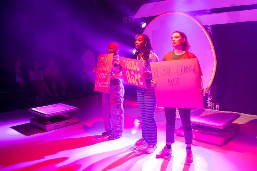 Three people stand on stage, each holding a sign. The signs read Our bodies our choice, There is hope, and People change not climate change. Purple and pink stage lighting creates a vibrant atmosphere. Audience members are visible in the background.