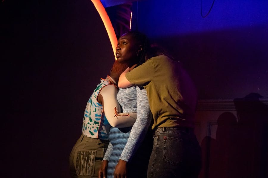The character Jamie (a young black woman with dark, braided hair, wearing a grey top and patterned blue leggings) looks out over the stage in disbelief. She is hugged tightly by two other people.