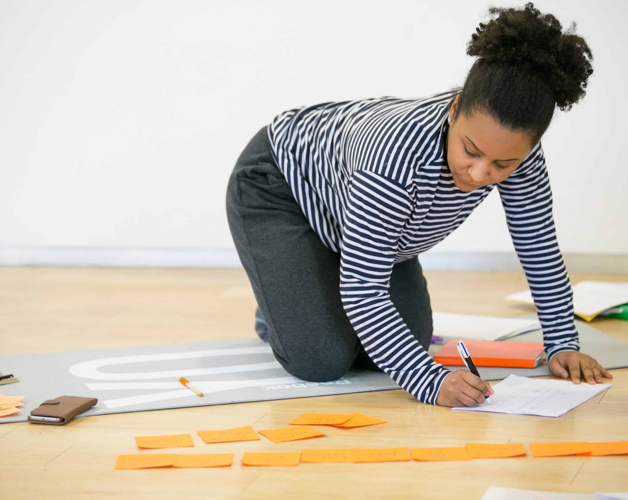 A person with curly hair, wearing a striped shirt and dark pants, kneels on a mat while writing on paper. Orange sticky notes and a smartphone are placed on the floor nearby. The background is a plain wall.