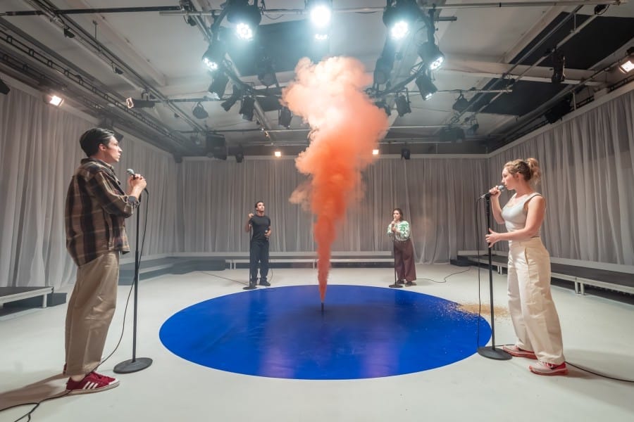 Four performers stand at microphone stands around a blue circle painted on the floor with a smoke flare going off in the middle of the circle