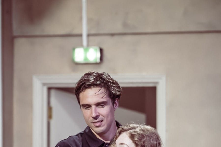 Ziggy stands behind Harriet with his hands resting on her shoulders