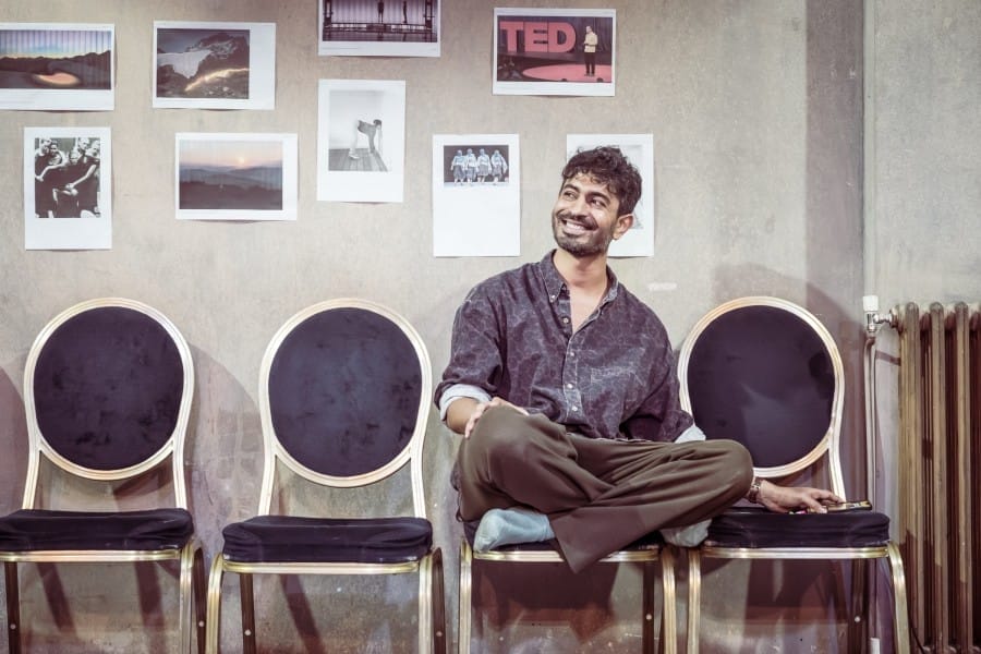 A smiling man sits cross-legged on the middle chair of a row of chairs against a wall.