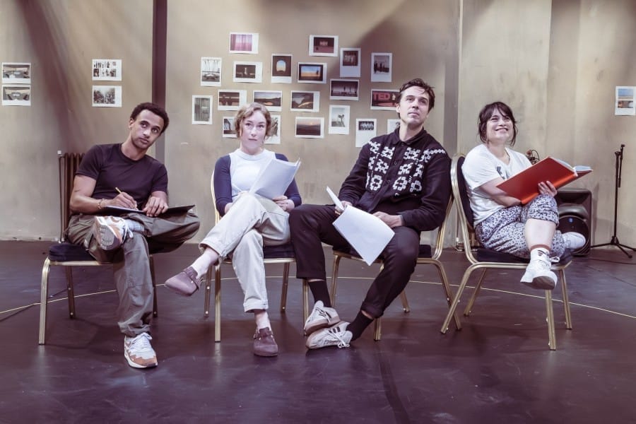 Four people sitting in a row on chairs, looking at the camera and holding scripts.