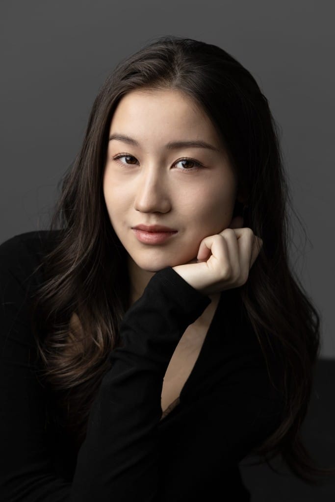 Naomi, a brunette East Asian woman in a black shirt has her hand up to her chin and is looking down the camera against a grey backdrop.
