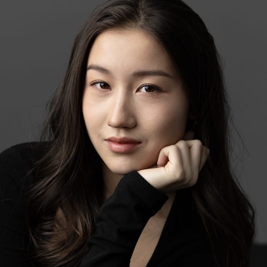Naomi, a brunette East Asian woman in a black shirt has her hand up to her chin and is looking down the camera against a grey backdrop.